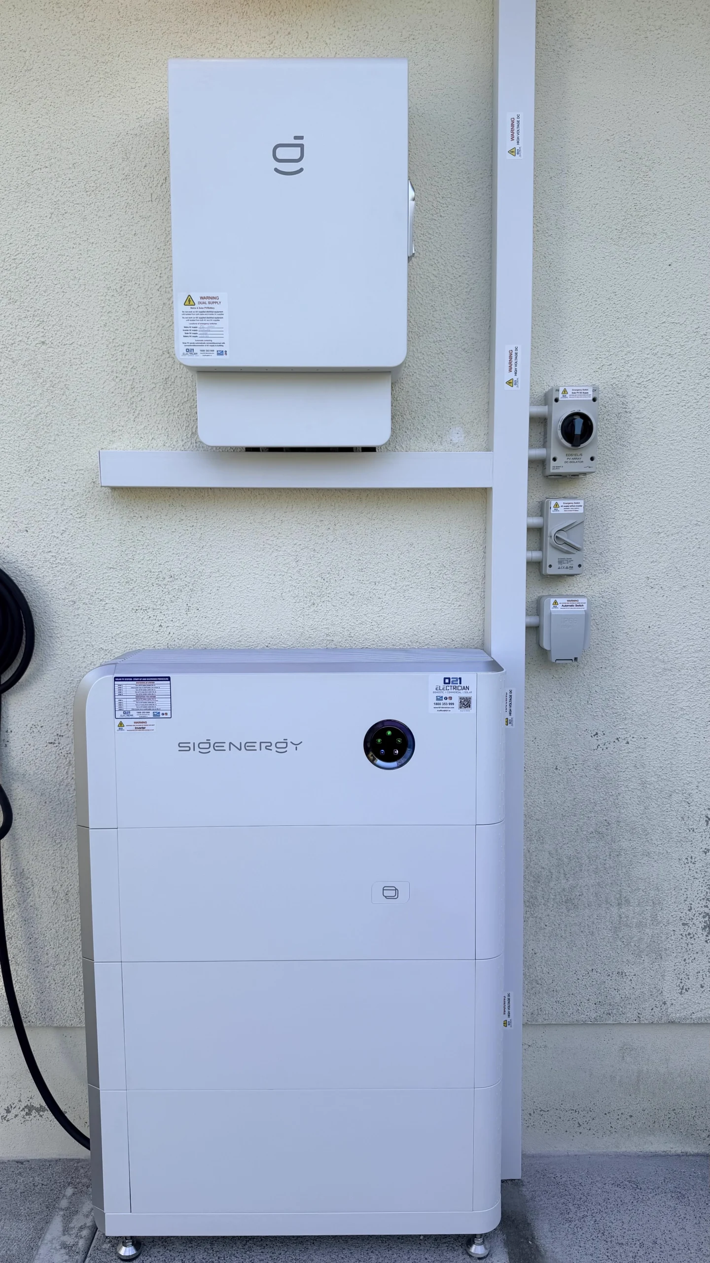 Wall-mounted Sigenergy solar inverter and multi-stack floor-standing battery storage system installed outdoors against a rendered wall, complete with protective white trunking and safety isolator switches.