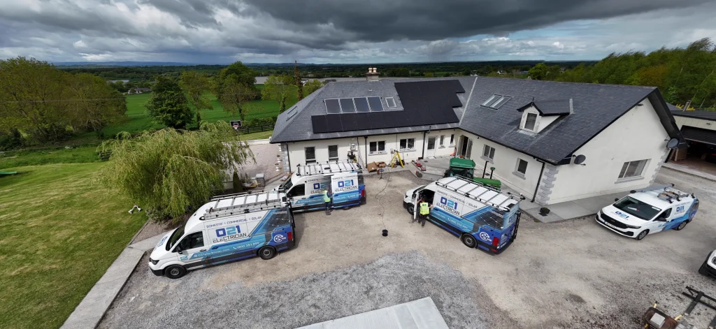 Aerial view of a residential property featuring an extensive rooftop solar panel array, with multiple branded 021 Electrician and 021 Solar service vans parked in the driveway below.